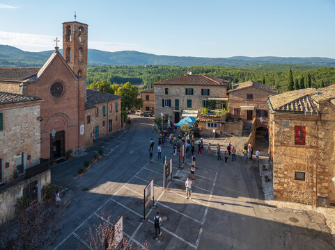 Aerial view of the Piazza di Ponte allo Spino bathed in warm sunlight, revealing the church and gathering crowds against a backdrop of rolling hills, Sovicille, Toscana, Italy.