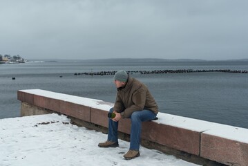 A young man holding a mug sits on a rocky embankment by the water. Winter pastime in the city.