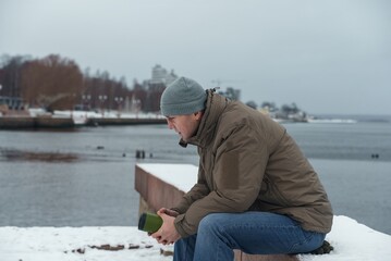A young man enjoys relaxing while sitting on a snow-covered rocky embankment. A walk by the water in winter. Outdoor recreation in winter. Side view.
