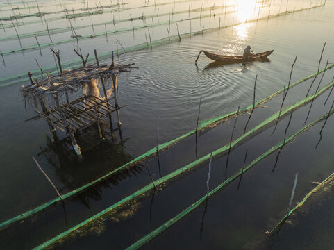 Aerial view of a lone figure in a small boat navigates through a grid of nets surrounding a weathered wooden structure, shimmering in the golden light of dusk, Tuy An, Phu Yen, Vietnam