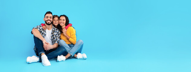 Three friends relax on a blue backdrop, sharing smiles and laughter. Two adults sit close together while a child embraces them, creating a joyful atmosphere.