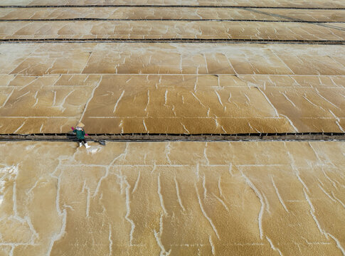 Aerial view of a lone worker raking across vast salt fields, the earthy tones contrasting with crisp white salt, Phuoc Diem, Ninh Thuan, Vietnam.