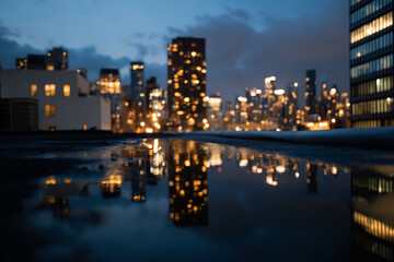 Obraz premium A captivating cityscape at twilight, with luminous skyscrapers reflected in rainwater puddles, symbolizing the beauty and charm of urban life amidst weather changes.