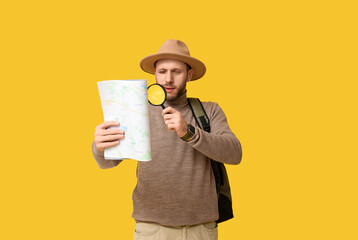 Young male tourist with magnifying glass looking at map on yellow background. Itinerary concept
