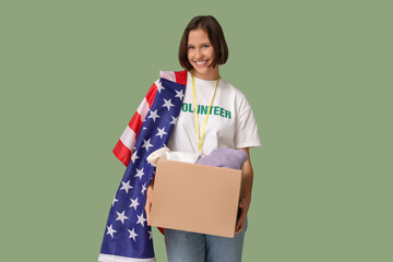 Young female volunteer holding USA flag and cardboard box with clothes on green background