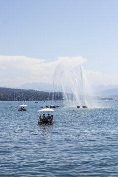 View of pedal boats gliding across the lake's surface, a grand fountain spraying high against a backdrop of distant mountains, Zurich, Switzerland.