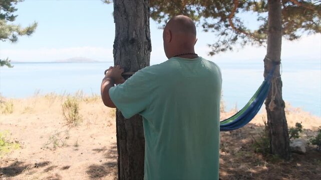 An adult bald man uses a camping knife to make notches on the tree trunk so the hammock holds firmly and does not slip down. He secures the hammock tightly for rest on the seaside.