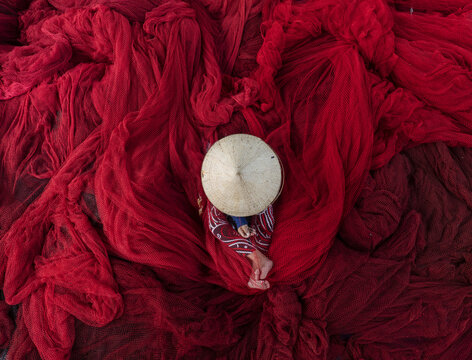 Aerial view of a person in a conical hat sits amidst a sea of vibrant red fishing nets, creating a striking contrast of color and texture, Thuan An, B&igrave;nh Duong, Vietnam.