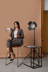 Young happy female film director with megaphone and studio lighting sitting on chair against beige...