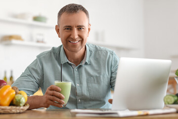 Mature man with glass of fresh vegetable smoothie using laptop in kitchen
