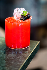 A vibrant red cocktail in a rocks glass, garnished with crushed ice, fresh blackberries, and a mint sprig on a green marble counter.
