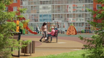 Caucasian Friends Share Quiet Playground Moment On Bench Under Trees, Apartment Facades And Colorful Play Equipment Visible, Relaxed Leisure And Soft Green Framing, Gentle Conversation And Shared