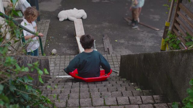 Boy sliding down outdoor stone staircase in bright red sled with excited anticipation turning into lively movement while siblings watch below creating playful adventurous energetic childhood moment