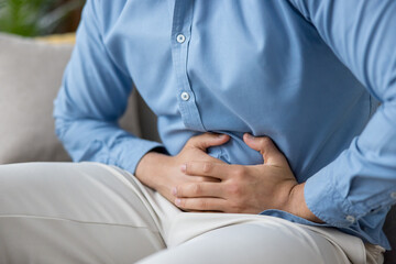 Obraz premium Man clutching his abdomen in visible discomfort from indigestion or cramps, seated indoors in casual clothes, close-up showing pain, illness, and digestive distress
