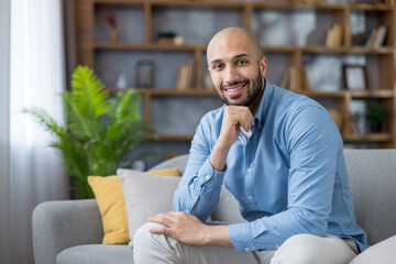 Bald, bearded man sitting confidently on a modern sofa in his cozy living room, smiling at the...