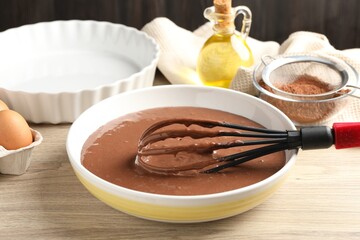 Liquid chocolate dough in bowl, whisk, oil, cocoa powder and egg on wooden table, closeup