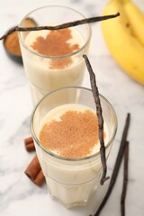 Delicious protein shake in glasses, bananas, cinnamon and vanilla pods on white marble table, closeup