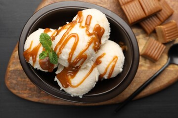 Tasty ice cream with caramel sauce and mint in bowl on black table, flat lay