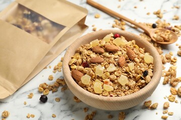 Tasty granola with dried fruits and nuts in bowl and paper bag on white marble table, closeup