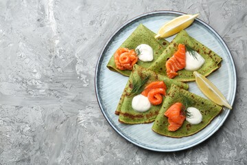 Tasty spinach crepes with salmon, sour cream, dill and lemon on grey table, top view. Space for text