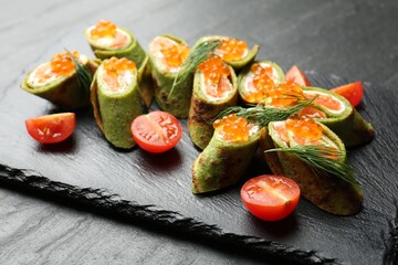 Tasty spinach crepes with salmon, cream cheese, red caviar, tomatoes and dill on black table, closeup