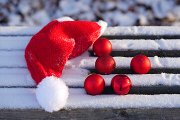 Red Christmas hat resting on a snowy wooden bench beside shiny red ornaments, creating a festive winter scene filled with holiday cheer and seasonal spirit