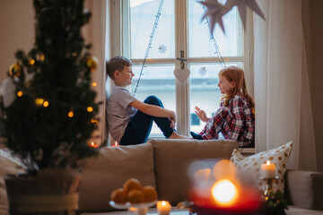Small girl and boy sitting on window sill in pajamas. Christmas time.