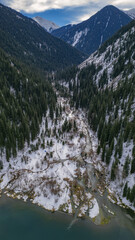 Drone shot of a snowy mountain valley surrounded by dense pine forests and rugged peaks. A winding river flows through the winter landscape, creating a dramatic and peaceful alpine scene.