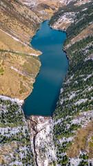 A top-down drone shot of a deep blue mountain lake surrounded by rugged alpine slopes. Snow patches, evergreen forests, and rocky terrain frame the winding river flowing into the lake. A striking high