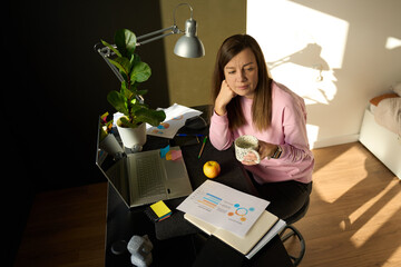 Woman sitting at home desk holding mug next to laptop, documents and office supplies