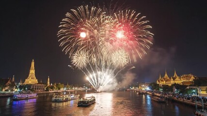 Vibrant fireworks light the night sky above a river, reflecting on the water. Illuminated traditional buildings and festive boats create a grand spectacle. Crowds watch, making this ideal for celebrat