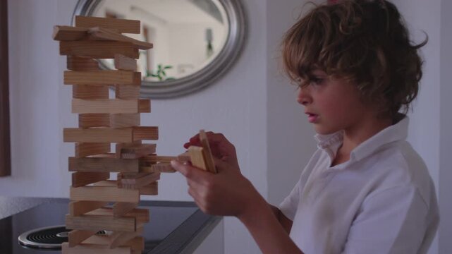 Boy adjusting wooden pieces on intricate tall tower with focused steady movements while shaping balanced structure during calm creative playtime inside bright relaxed home environment