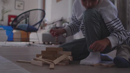 Boy building wooden block structure on floor as off-camera piece suddenly lands and disrupts his careful work creating surprise reaction and playful chaotic moment in cozy family home