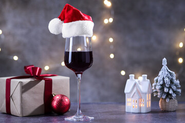 Glass of red wine with Santa hat and Christmas decor on grey table against background with blurred lights