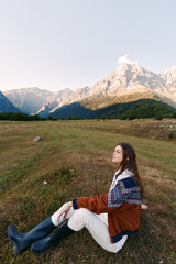 Fototapeta premium Woman sitting in a meadow wearing a patterned sweater and rubber boots, enjoying mountains and open field at golden hour, serene outdoor portrait and travel moment in nature