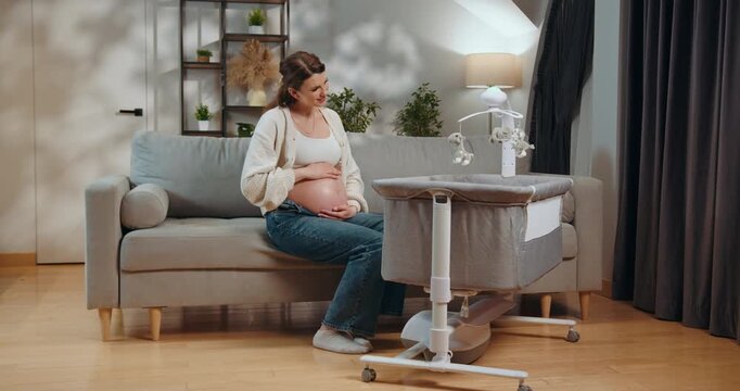 Pregnant woman sits on sofa touching belly near bedside bassinet with mobile. Pregnant Woman With Bassinet