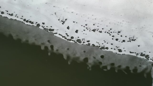 The edge of a river that has defrosted just enough to show the water flowing under the ice and snow layer. The melting snow leaves a unique pattern in the ice on top. 4k, drone, pulling up and away
