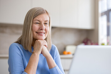 Fototapeta premium Senior woman smiling with hands clasped, listening and connecting on a video call using a laptop in her kitchen, embracing technology for communication