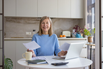 Smiling mature woman at her kitchen table using a laptop and paperwork to manage household...