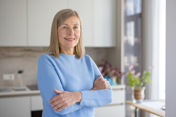Senior woman standing with crossed arms in a bright modern kitchen, looking directly at the camera...