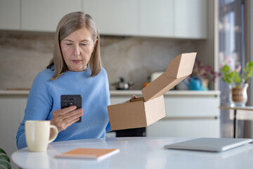 Senior woman at home unboxing a parcel, holding her smartphone, comparing contents with an online...