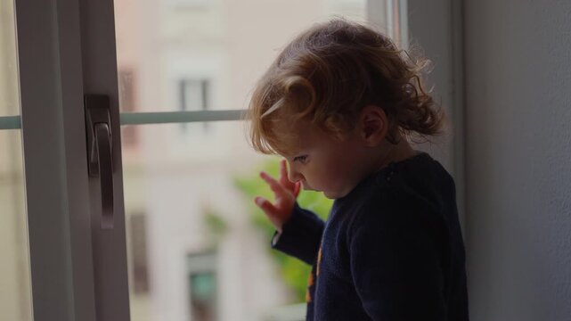 Toddler reaching toward window handle without being able to open it while standing indoors near closed frame illustrating protective home safety measures around accessible window
