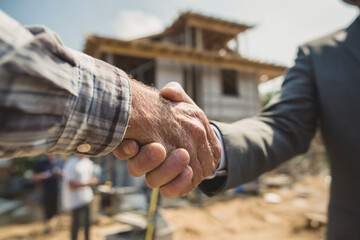 Close-up of a construction worker in a checkered shirt shaking hands with a businessman in a suit on a residential building site, symbolizing agreement and real estate partnership.