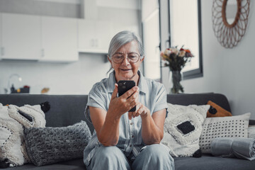 Elderly woman sits on a sofa at home using her smartphone to browse and shop online. She focuses on...