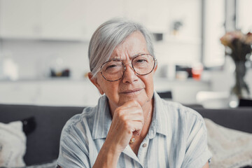 Elderly woman sits on a sofa at home looking at the camera with a suspicious expression. She appears concerned about online security and potential scams while using her devices.