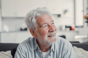 Elderly man sits on a sofa at home looking outside the window. He feels calm and reflective while...