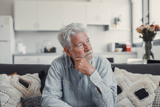 Senior man sits alone on a sofa looking outside with a sad expression. He seems stressed and reflective while dealing with personal concerns at home.
