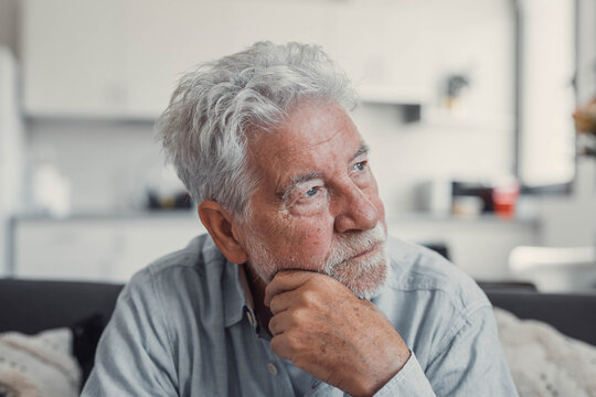 Senior man sits alone on a sofa looking outside with a sad expression. He seems stressed and reflective while dealing with personal concerns at home.