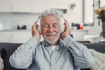 Elderly man sits on a sofa at home enjoying music with headphones. He feels relaxed, happy, and...