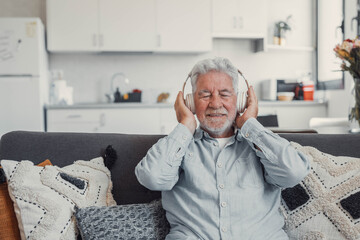 Elderly man sits on a sofa at home enjoying music with headphones. He feels relaxed, happy, and...